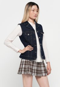 Denim prsluk tamnog pranja, strukturiranog dizajna, s prednjim džepovima i srebrnim dugmadi. Uparen s laganom bluzom i plisiranom suknjom kariranog uzorka.