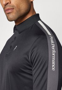 Polo shirt nera con colletto, caratterizzata da una striscia grigia chiaro e un logo sulla manica. Realizzata in un tessuto liscio e testurizzato.