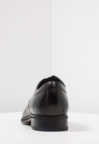 Zapato de vestir de cuero negro con textura lisa, punta redondeada, tacón bajo y una sutil marca grabada en el tacón.