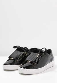 Schwarze Lackleder-Sneaker mit weißen Sohlen, schwarzen Schnürsenkeln und dekorativem schwarzen Fransen über den Schnürsenkeln, auf einer weißen Fläche dargestellt.