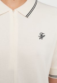 Polo crema con cierre de cremallera, cuello a rayas en blanco y negro, tejido texturizado y un logo bordado en negro.