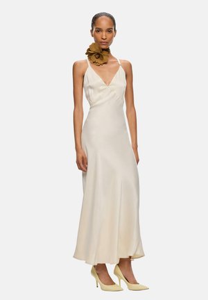 Mujer que lleva un vestido sin mangas color crema con un escote en V profundo, un accesorio para el cuello con flores marrones y tacones altos puntiagudos en color crema.