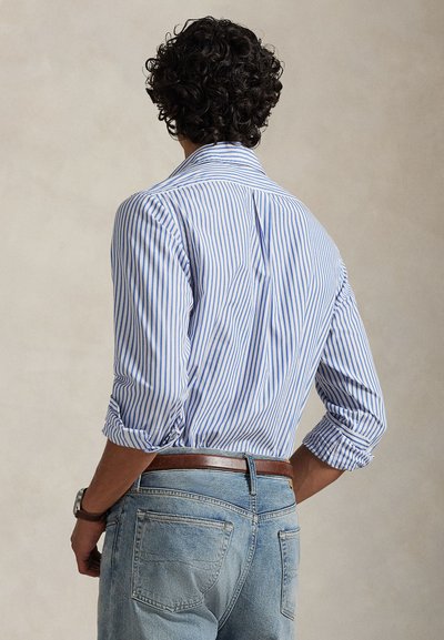 Μπλε και λευκό ριγέ πουκάμισο από ύφασμα με μανίκια που έχουν γυριστεί προς τα πάνω· φορεμένο με ανοιχτόχρωμα τζιν και καφέ ζώνη.