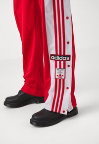 Calças desportivas vermelhas com detalhes em riscas brancas, apresentando botões de pressão, remendos do logo preto da Adidas e usadas com sapatos pretos.