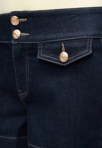 Λεπτομέρεια σκοτεινής μπλε τζιν φούστας με χρυσά διακοσμητικά κουμπιά και ραφές αντίθεσης σε καφέ απόχρωση στη μέση και την τσεπάκι.