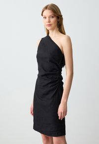 Abito nero asimmetrico realizzato in un tessuto testurizzato, con dettagli arricciati sui lati e una silhouette aderente. Il design a spalla unica mette in risalto la semplicità.