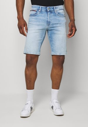 Homme portant un short en jean bleu clair, des chaussettes blanches montantes et des baskets blanches avec des logos étoile noirs, debout devant un fond uni.