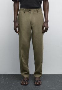 Pantaloni in lino verde oliva con vestibilità dritta, tasche frontali e chiusura con un singolo bottone nero, abbinati a infradito marroni.