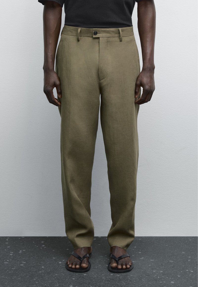 Pantaloni in lino verde oliva con vestibilità dritta, tasche frontali e chiusura con un singolo bottone nero, abbinati a infradito marroni.