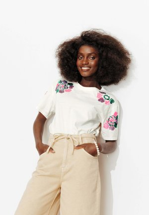 Femme aux cheveux bouclés et volumineux sourit, portant une chemise blanche à manches courtes avec une broderie florale rose et verte et un pantalon beige taille haute.