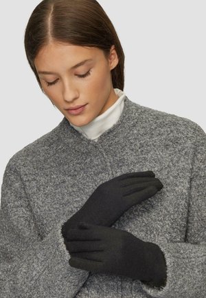 Donna che indossa guanti neri e un cappotto grigio texturato, aggiustando un guanto con le mani e guardando in basso.