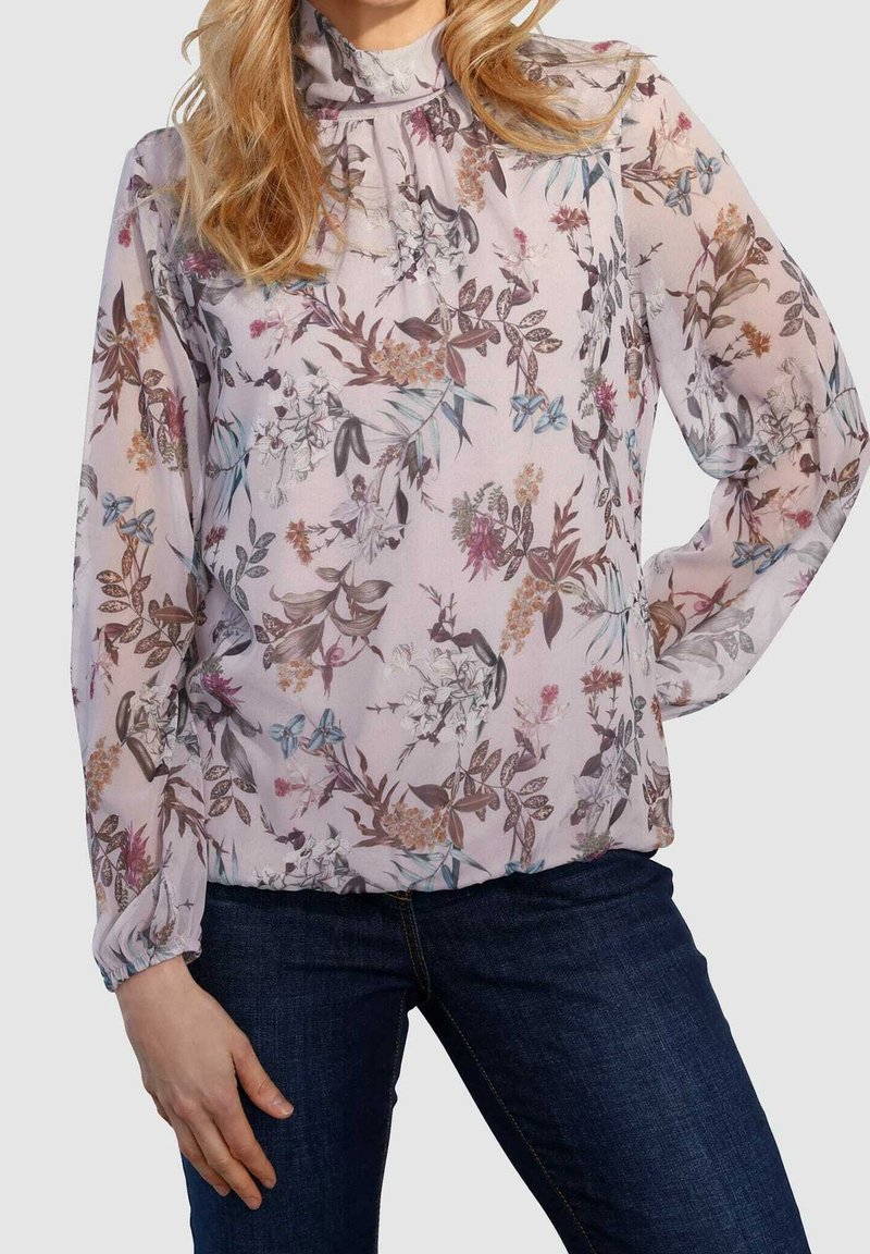 Blusa fluida realizzata in tessuto trasparente, con uno sfondo rosa chiaro e motivi floreali in viola, blu e marrone. Maniche lunghe con polsini arricciati.
