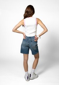 Hvit ermeløs topp med rosa bølgete detaljer, paret med blå denimshorts og hvite joggesko med lilla detaljer, sett fra bak.