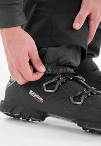 Svart skidstövel med texturerad yta, metallkomponenter och röda accenter. Handjusterar elastiken på benen av svarta vinterbyxor.