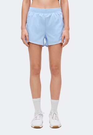 Persoană purtând pantaloni scurți sportivi bleu deschis, șosete albe și pantofi de alergare albi, stând în fața unui fundal simplu.