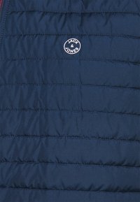 Granatowa kamizelka puchowa z poziomymi pikowanymi liniami, miękką teksturą oraz okrągłą naszywką z logo "JACK & JONES" w białym kolorze na przedzie.