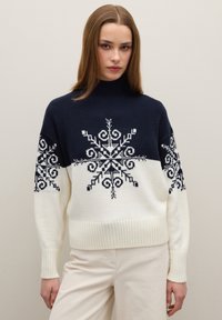 Pull en tricot bleu marine et crème, avec des motifs flocons de neige noirs. Col montant, poignets et ourlet côtelés, coupe décontractée. Texture douce.