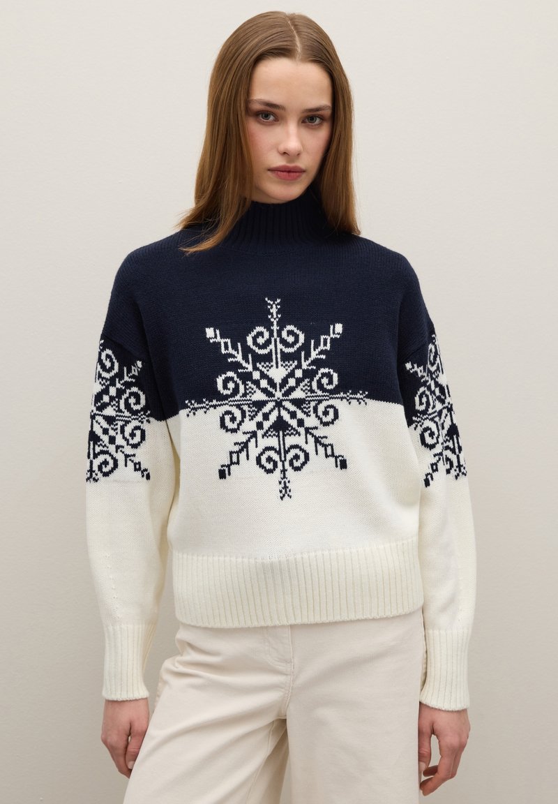 Pull en tricot bleu marine et crème, avec des motifs flocons de neige noirs. Col montant, poignets et ourlet côtelés, coupe décontractée. Texture douce.