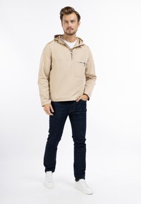 Lichtbeige, met capuchon, pulloverjack met een voorzak met rits. Zachte, gladde textuur; gedragen over donkerblauwe jeans en witte sneakers.
