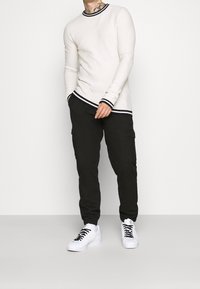 Žebrovaný bílý top s dlouhým rukávem a černými akcenty, v kombinaci s černými cargo kalhotami a bílými teniskami s černými tkaničkami.