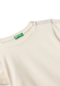 Krémové tričko s krátkým rukávem a kulatým výstřihem se zelenou nálepkou United Colors of Benetton na límci.