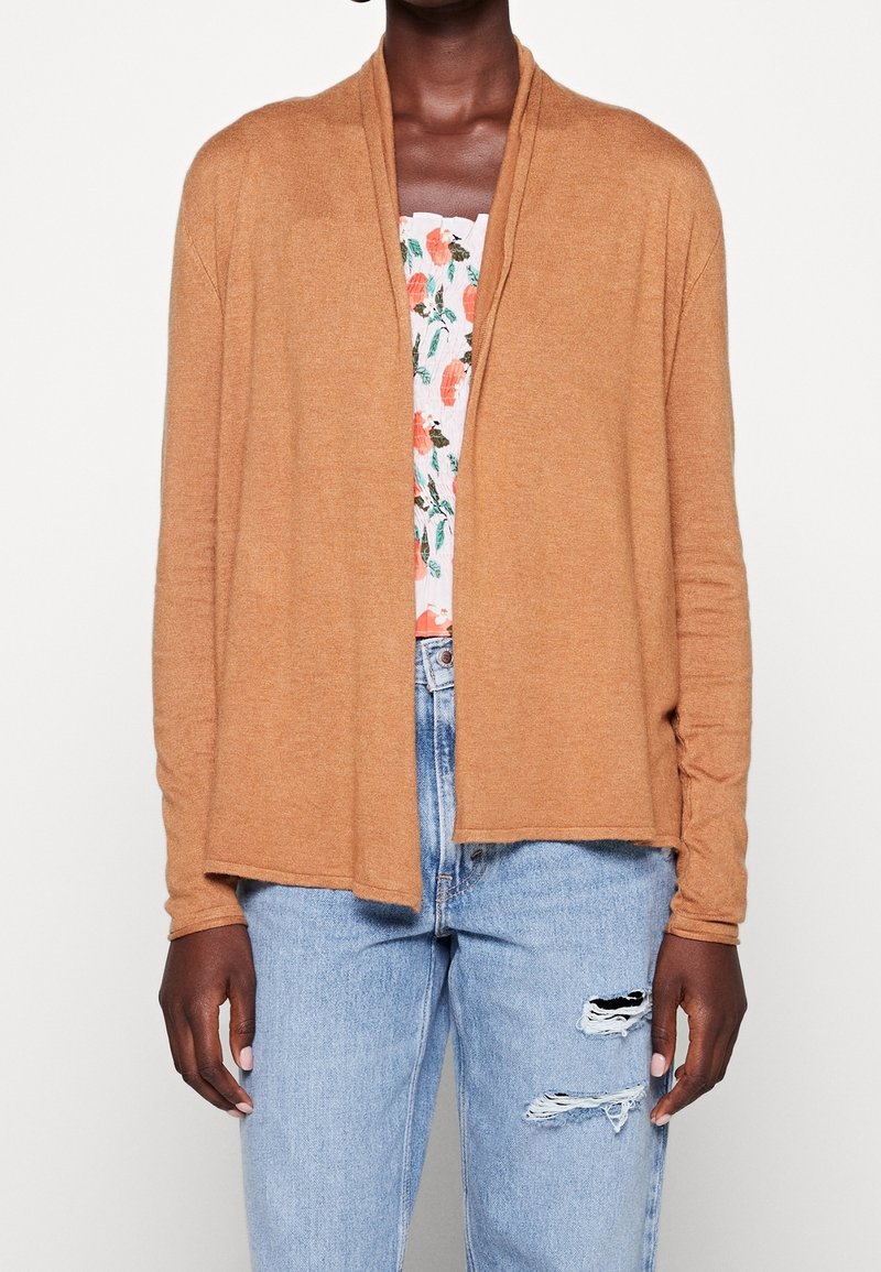 Personne à la peau foncée portant un cardigan ouvert marron clair, un haut à fleurs et un jean bleu clair déchiré, debout devant un fond blanc.
