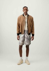 Homem de pé, vestindo uma jaqueta bomber castanha, camisa branca, calções cinzentos de camuflagem, meias brancas e ténis bege, em um fundo simples.