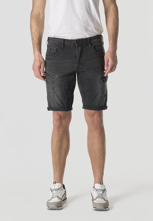 Hombre con pantalones cortos cargo negros remangados, camiseta blanca y zapatillas blancas, de pie frente a un fondo liso.