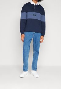 Polo de manga larga en azul marino y gris con cuello blanco, que presenta un logo en el pecho, combinado con jeans rectos de pierna ligera en azul claro y zapatillas blancas.