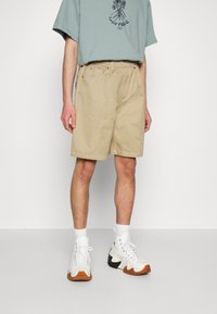 Pantaloni beige con vestibilità rilassata, passanti per cintura, tasche frontali e dettagli di cucitura laterale. Abbinati a una maglietta grafica blu chiaro e scarpe da ginnastica bianche.