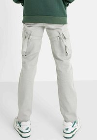 Cargo broek in lichtgrijze stof met meerdere zijzakken en verstelbare banden bij de taille. Draag met witte en groene sneakers.