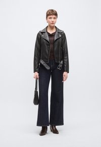 Μαύρο δερμάτινο μπουφάν μοτοσυκλέτας, καφέ τοπ, σκοτεινό τζιν με φαρδιά μπατζάκια και καφέ μποτάκια μέχρι τον αστράγαλο. Το μοντέλο κρατά μια μικρή μαύρη τσάντα χειρός.