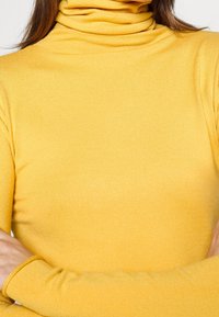 Maglione giallo a collo alto realizzato in tessuto a coste. Caratterizzato da maniche lunghe e una vestibilità aderente, con una texture morbida e un collo alto.