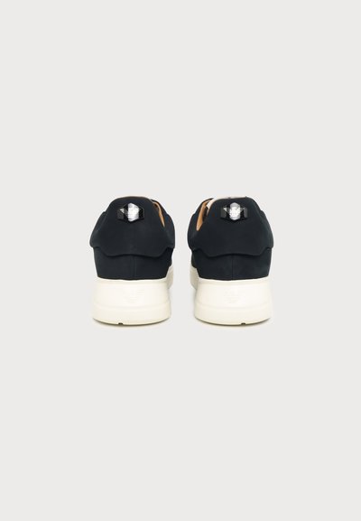 Paire de baskets noires avec semelles blanches vues de dos sur un fond clair uni, montrant les logos des marques sur les talons.