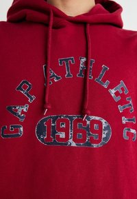 Rode hoodie van katoenmix, met een voorkantgrafiek waarin in navyblauwe, versleten letters "GAP ATHLETIC" en "1969" staat.