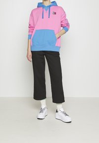Felpa con cappuccio colorata in rosa e blu, dotata di tasca frontale, abbinata a pantaloni cropped neri e sneakers bianche.
