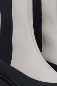 Bílé kožené kotníkové boty mají černou žebrovanou látkovou vložku, zaoblenou špičku a tlustou černou podrážku, což ukazuje moderní, strukturovaný design.