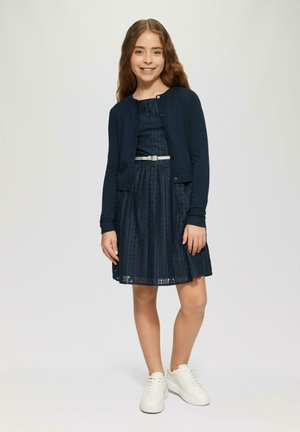 Jūras spēku zilo adīto kardigānu virs vairākkārtīgā zila kleita ar rakstainu tekstūru, izceltu jostasvietu ar sudraba jostu, kombinējot ar baltām kedām.