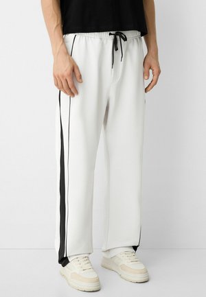 White athletic pants with black side stripes, elastic waistband, and drawstring. Smooth fabric with a relaxed fit. Paired with beige sneakers.