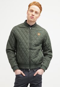 Jachetă verde matlasată cu fermoar frontal, cu manșete și tiv din tricot. Include un patch cu logo și buzunare laterale, realizată dintr-un material sintetic neted.