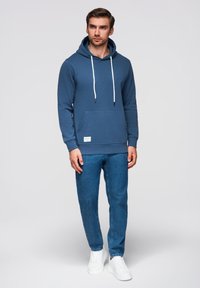 Felpa blu con cappuccio, lacci bianchi, tasca frontale; abbinata a jeans azzurri chiari e sneakers bianche. Texture liscia e design casual.