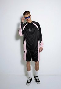 Czarny sportowy sweter i szorty z różowymi akcentami oraz prążkowaną teksturą. Model nosi niebieskie okulary przeciwsłoneczne, białe skarpetki i czarne sneakersy.