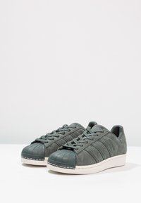 Zapatillas de ante grises con punta de goma, superficie texturizada, seis agujeros para los cordones y franjas laterales sutiles. Media suela blanca con detalle de costura.