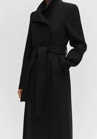 Cappotto nero in misto lana con maniche lunghe, colletto alto, chiusura a doppio petto con bottoni e una cintura coordinata annodata in vita.
