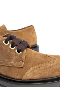 Chaussures en daim marron avec une texture lisse, design à bout rond, œillets en métal et lacets marron foncé. Présentent un léger talon et des coutures contrastées.