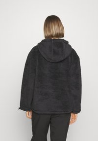Schwarze Fleece-Jacke mit Kapuze, ausgestattet mit einer weichen Textur, weit geschnitten und mit elastischen Zugbändern am Saum. Nahtdetail auf dem Rücken.