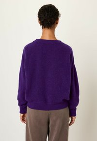 Maglione viola oversize e peloso con scollatura a girocollo e polsini a coste. Abbinato a pantaloni marroni, larghi e comodi. La texture morbida è ben visibile.