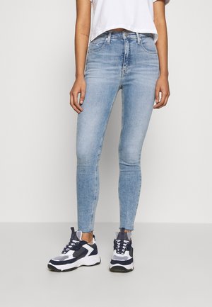 Person iført lyseblå skinny jeans, hvid cropped t-shirt og sort-hvide chunky sneakers står mod en ensfarvet baggrund.