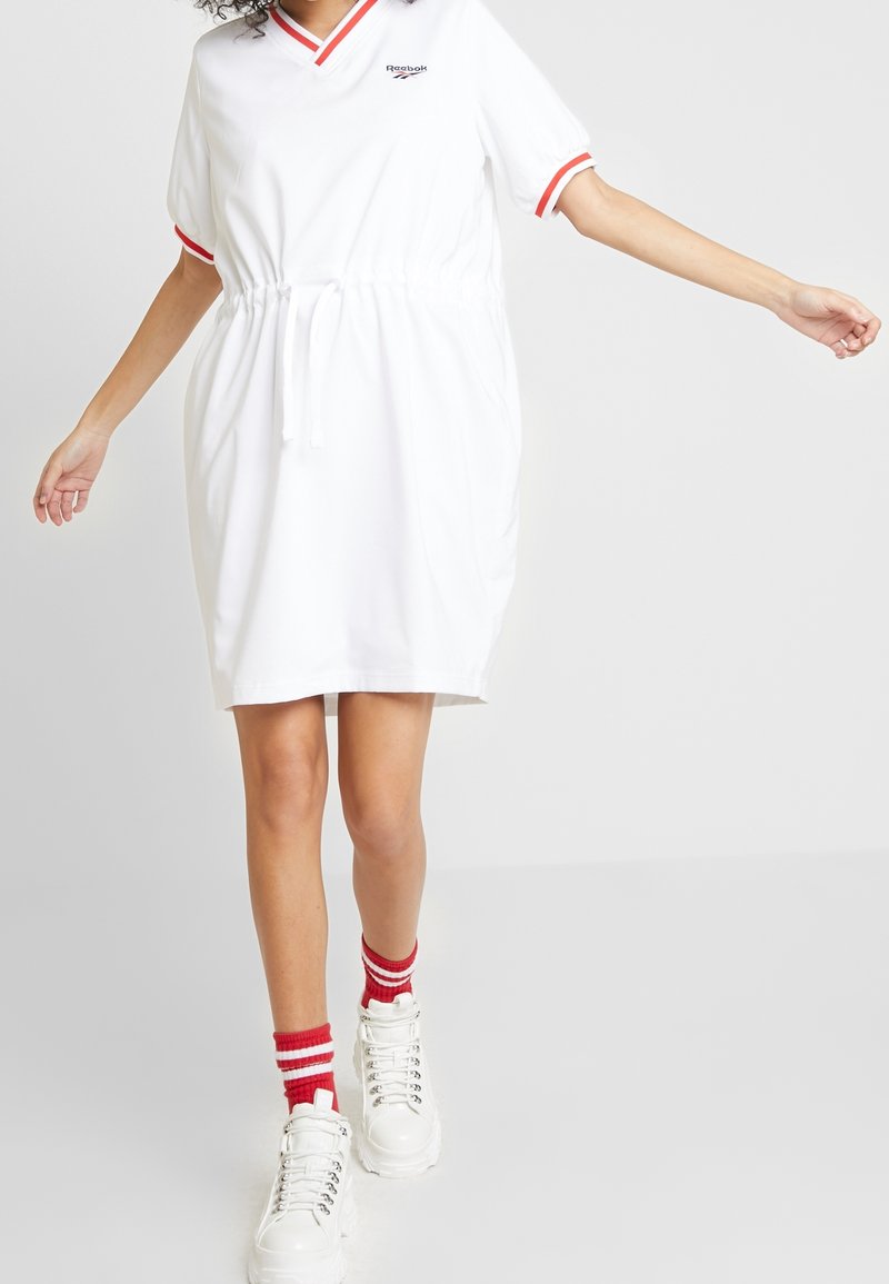 Femme portant une robe blanche Reebok avec des finitions rouges, des chaussettes rayées rouges et des baskets blanches à plateforme, marchant avec les bras légèrement étendus.