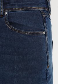 Jeans de mezclilla azul oscuro con costuras naranja, que cuentan con un bolsillo frontal con un remache de metal y una textura suave sin desgaste visible.